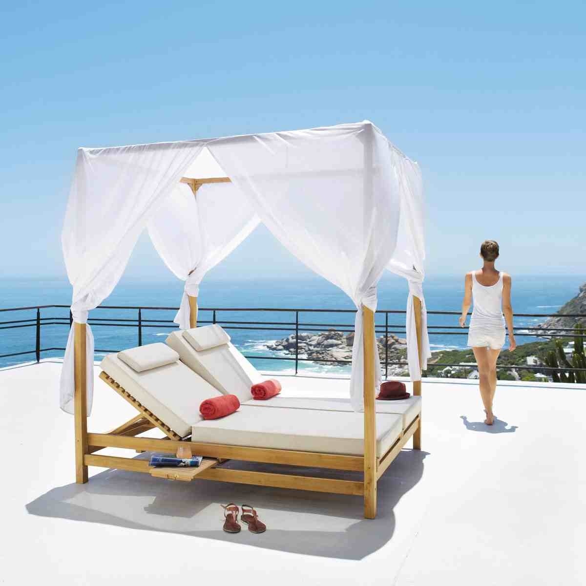 a four poster sunbed with voile curtains sits on a balcony overlooking a blue sea and sky. A brunette stands looking out to sea with her back to the camera she is slim and wearing a wihte vest top and white shorts. Her sandals are next to the sunbed.